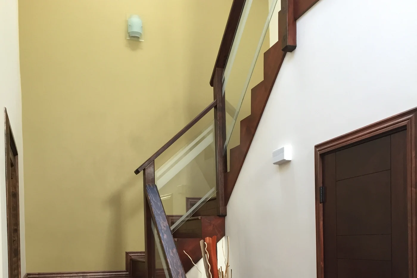 A stairwell where a soft yellow wall meets a white one, with a stained wood handrail, glass panels and a dark stained door