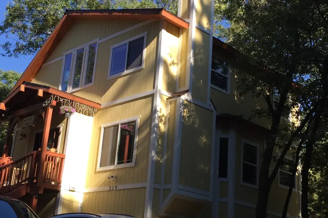 Three-story mountain home painted pale yellow with white trim, an orange-stained gable and a redwood porch